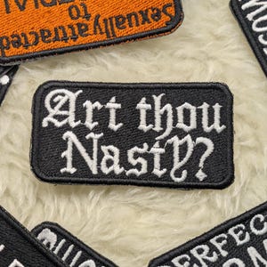 Può includere: Toppa ricamata nera con la frase "Art thou Nasty?" in lettere gotiche bianche. La toppa è rettangolare con angoli arrotondati. Altre toppe con testo sono visibili sullo sfondo.