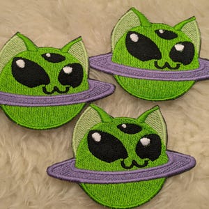 Könnte beinhalten: Drei grüne Alien-Katzen-Stickpatches mit drei Augen und einem lila Planetenring. Die Alien-Katzen haben schwarze Augen und weiße Pupillen.