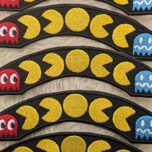Puede incluir: Parches bordados con fondo negro, personajes amarillos de Pac-Man, un fantasma rojo y un fantasma azul. Los parches son arqueados y tienen un tema de videojuego retro.