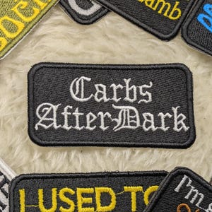 Peut inclure: Patch brodé noir et blanc avec le texte "Carbs After Dark" dans une police gothique.