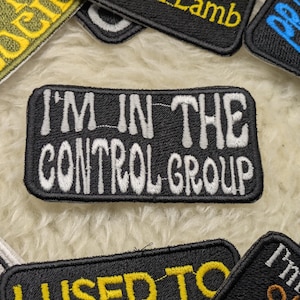 May include: A black rectangular patch with white embroidered text reading "I'M IN THE CONTROL GROUP." Other patches with different text and colours surround it. These patches are likely for clothing or accessories.