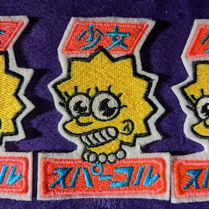 May include: Three embroidered patches featuring a yellow cartoon character with large eyes and a toothy grin. The character is wearing a pearl necklace and has a pink and blue banner above its head that reads "少女". The bottom banner reads "スーパーコル".