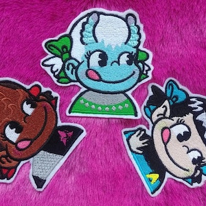 May include: Three embroidered iron-on patches featuring cartoon characters. The first patch is a brown-skinned girl with a red bow. The second patch is a blue-skinned girl with green hair and a green bow. The third patch is a black-haired girl with a blue bow.