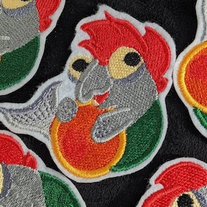 May include: Embroidered iron-on patch featuring a cartoon fish with red hair, holding a red and orange life preserver. The fish is gray with green and white accents.