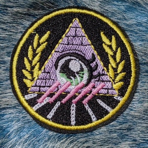 May include: A black and gold embroidered patch with a purple pyramid, an eye, and a wreath. The pyramid has pink stripes and the eye is surrounded by a black circle.