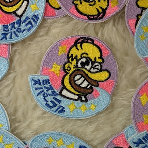 May include: Embroidered patch featuring a yellow cartoon character with a large grin, set against a pink and purple background. The patch includes blue accents with Japanese text and gold stars, perfect for adding to clothing or accessories.