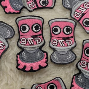 Peut inclure: Patchs en forme de bombe de peinture rose et gris avec le mot "977D" dessus. Les patchs ont des yeux blancs et une étoile noire au milieu de chaque œil.