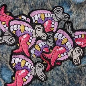 Könnte beinhalten: Ein Set mit sechs gestickten Patches, die einen offenen Mund mit Zähnen und einer Zunge in Lila und Pink zeigen. Die Patches haben jeweils ein graues mechanisches Detail.