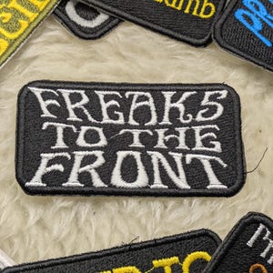 Könnte beinhalten: Schwarzer gestickter Aufnäher mit weißem Text, der "FREAKS TO THE FRONT" lautet.
