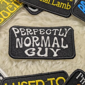 Könnte beinhalten: Schwarzer gestickter Aufnäher mit weißem Text, der "Perfectly Normal Guy" lautet.