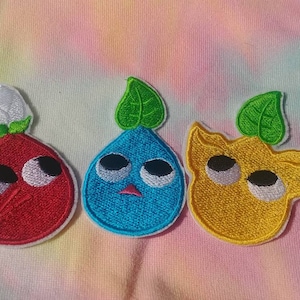 May include: Three embroidered iron-on patches featuring cartoon characters with large eyes. The characters are a red, a blue, and a yellow, each with a green leaf on top.