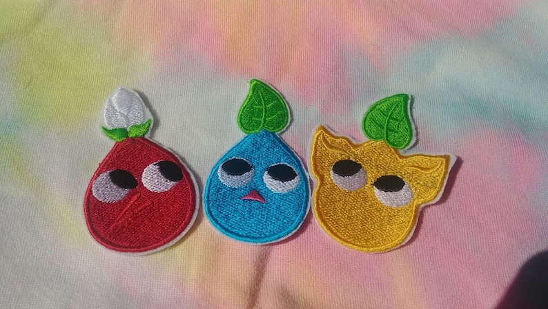 May include: Three embroidered iron-on patches featuring cartoon characters with large eyes. The characters are a red, a blue, and a yellow, each with a green leaf on top.