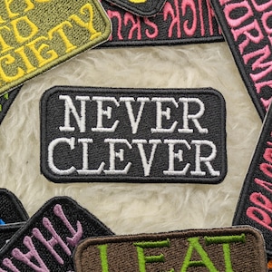May include: A black and white embroidered patch with the text "NEVER CLEVER" in white lettering. The patch is surrounded by other colourful patches with various text.