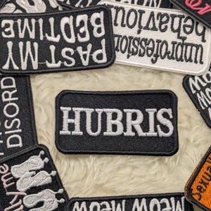 Puede incluir: Un conjunto de parches bordados de varias formas y tamaños. Los parches son negros, blancos y naranjas, con frases como "HUBRIS", "PAST MY BEDTIME" y "MEOW MEOW" bordadas en blanco. Los parches están dispersos sobre una superficie suave y blanquecina.