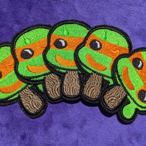 May include: Five green, orange and black embroidered patches of Teenage Mutant Ninja Turtles. Each patch features a different turtle with a unique expression.