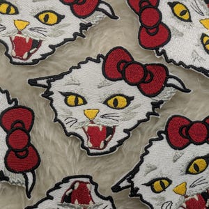 Puede incluir: Parches bordados con la cara de un gato blanco con ojos amarillos, boca roja abierta y un lazo rojo. Los parches tienen un contorno negro y están dispersos sobre una superficie texturizada.