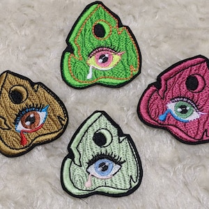 May include: Four embroidered patches shaped like a house with a single eye and a tear. The patches are in different colors: green, gold, pink, and light green.