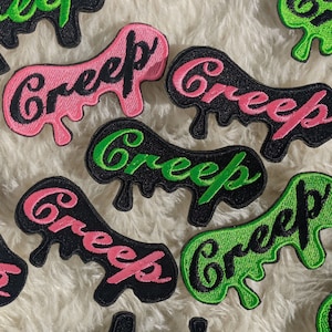 Op de afbeelding: Een verzameling strijkbare patches met het woord "Creep" geschreven in een druppelend lettertype. De patches zijn in verschillende kleuren, waaronder zwart, groen en roze.
