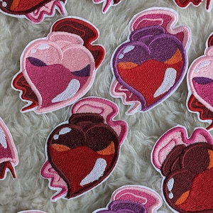 May include: A collection of embroidered iron-on patches shaped like hearts with a potion bottle design. The patches are in shades of pink, red, and purple with white accents.