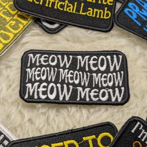 Può includere: Toppa ricamata in bianco e nero con il testo "MEOW MEOW MEOW MEOW MEOW MEOW" in lettere bianche su sfondo nero.