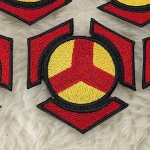 Puede incluir: Cinco parches bordados con un contorno negro y un diseño geométrico rojo y amarillo. Los parches están sobre una superficie blanca y peluda.