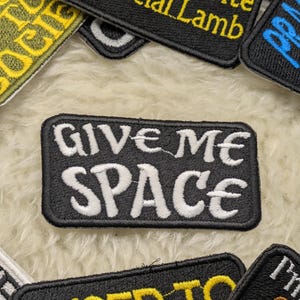 Peut inclure: Patch brodé noir et blanc avec le texte "Give Me Space" en lettres blanches sur un fond noir.