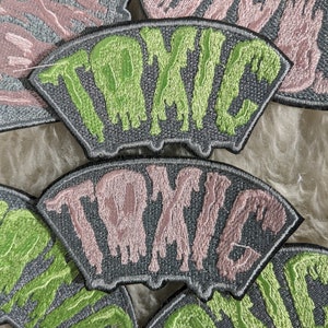 May include: Close-up of several embroidered patches with the word "TOXIC" in a dripping font. The patches are in various colors, including green, pink, and gray.