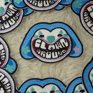 May include: Embroidered patches with a clown face design. The patches have a blue outline, a white face with a large grin, and the words "CLOWN GROUND" in a circular pattern. The patches are on a fluffy, white surface.