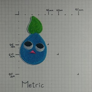 May include: A blue and green embroidered patch of a cartoon character with a single leaf on top. The character has large eyes, a pink mouth, and a blue body.