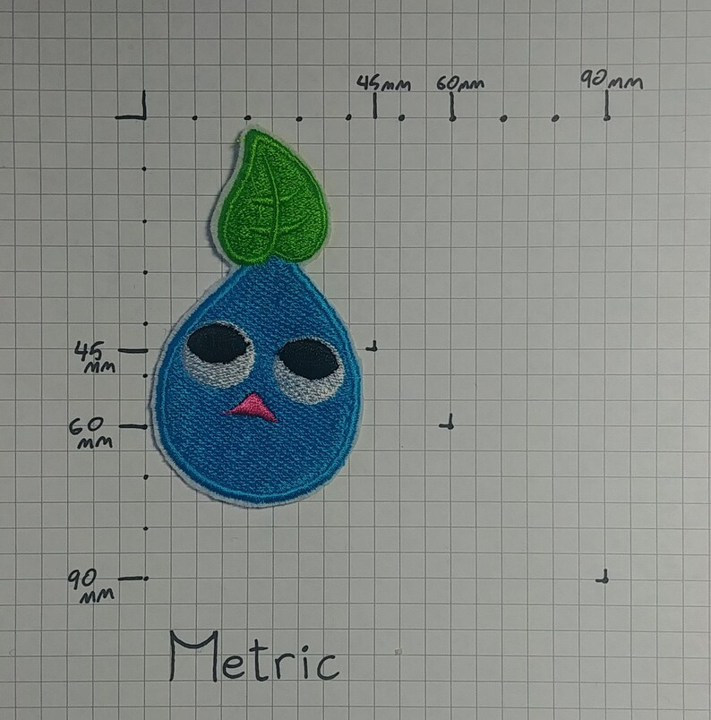 May include: A blue and green embroidered patch of a cartoon character with a single leaf on top. The character has large eyes, a pink mouth, and a blue body.