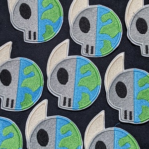 Könnte beinhalten: Gestickte Aufnäher mit einem Schädel-Design, das in zwei Hälften geteilt ist, eine Seite ein grauer Schädel und die andere eine blaue und grüne Erde. Jeder Aufnäher hat eine cremefarbene, flügelförmige Form. Dunkelblauer Hintergrund.