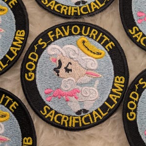 Könnte beinhalten: Ein schwarzer und weißer gestickter Aufnäher mit einem Comic-Lamm, das einen Heiligenschein und ein rosa Band trägt. Der Text "GOD'S FAVORITE SACRIFICIAL LAMB" ist in Gelb um das Lamm herum gestickt.