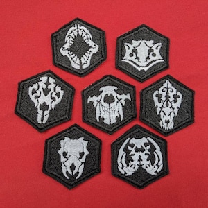 Peut inclure: Huit patchs hexagonaux noirs avec des motifs brodés blancs. Chaque patch présente un design de crâne stylisé différent.