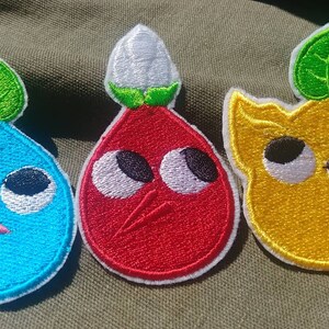 May include: Three embroidered patches featuring cartoon characters with large eyes. The characters are a blue teardrop-shaped character with a green leaf on top, a red teardrop-shaped character with a white flower on top, and a yellow character with a green leaf on top.