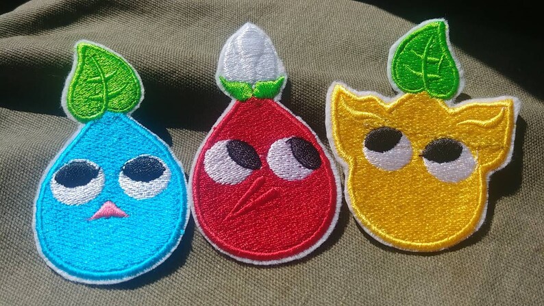 May include: Three embroidered patches featuring cartoon characters with large eyes. The characters are a blue teardrop-shaped character with a green leaf on top, a red teardrop-shaped character with a white flower on top, and a yellow character with a green leaf on top.