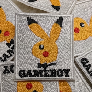 May include: A white embroidered patch featuring a yellow cartoon bunny wearing a black bow tie. The bunny has a red nose and black eyes. The text "GAMEBOY" is written in black below the bunny.