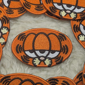 Peut inclure: Écussons Garfield brodés orange avec un contour marron et des traits du visage blancs et noirs. Les écussons sont de forme ovale et représentent le visage du chat de dessin animé. Pour vêtements ou accessoires.
