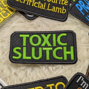 Może przedstawiać: Czarna łatka z białymi szwami i tekstem "TOXIC SLUTCH" w kolorze zielonym.