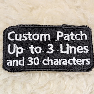 May include: Black rectangular patch with white embroidered text. The text reads "Custom Patch Up to 3 Lines and 30 characters". The patch has a black border and is displayed on a white, fluffy surface.