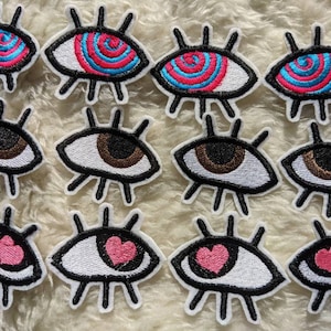 May include: Twelve embroidered iron-on patches featuring various eye designs. Some eyes have a swirl pattern in pink and blue, others have a brown iris, and some have a pink heart in the centre.
