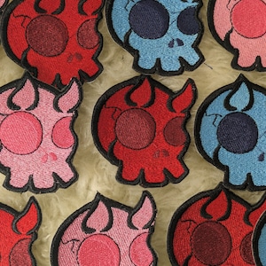 Puede incluir: Una colección de parches bordados con forma de calavera en varios colores, incluyendo rojo, rosa, azul y negro. Las calaveras tienen detalles en forma de llama alrededor de los bordes y están delineadas en negro.