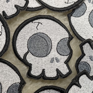 May include: A collection of embroidered iron-on patches featuring a cartoon skull design in shades of gray and white. The skull has a simple design with two large eye sockets and a small mouth.