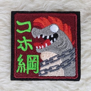 Puede incluir: Parche bordado con un monstruo de dibujos animados rojo, blanco y gris con una cadena alrededor del cuello. El monstruo tiene una boca roja con dientes afilados y una lengua verde. El texto "ゴキブリ" está bordado en verde en el parche.
