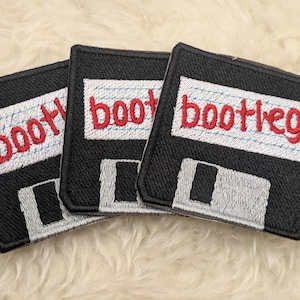 Puede incluir: Tres parches de tela negra con diseños de disquetes blancos y grises. Cada parche tiene la palabra "bootleg" escrita en bordado rojo.