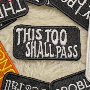 May include: A collection of embroidered patches with various phrases. The central patch is black with white text that reads "THIS TOO SHALL PASS." Other patches include the words "THREE," "unprofessional," and "PROBLEM." The patches are on a fluffy, off-white surface.