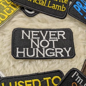 Op de afbeelding: Zwart-wit geborduurde patch met de tekst "NEVER NOT HUNGRY" in witte letters op een zwarte achtergrond.