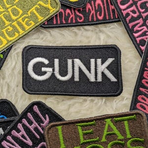 May include: A collection of embroidered patches with various phrases, including "GUNK", "EAT", "DRINK", "SOCIETY", and "REALIZING". The patches are black, green, pink, and yellow.
