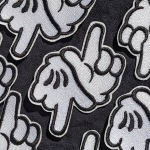 May include: Several white felt patches with black embroidered outlines of a hand giving the middle finger. The patches are on a black fabric background.