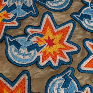 Puede incluir: Un conjunto de parches bordados para planchar que representan una explosión de dibujos animados con un rayo azul y blanco. Los parches son de varios colores, incluyendo azul, blanco, rojo, naranja y amarillo.