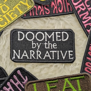 May include: A black and white embroidered patch with the text "Doomed by the Narrative". The patch is surrounded by other colourful embroidered patches with various text.